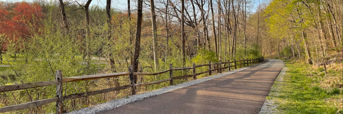 Paved walking path at Strubel Trail lined with a wooden split-rail fence and early spring trees just beginning to leaf out in shades of green and red — walking trails near West Chester