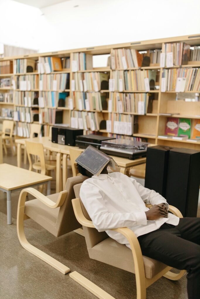 A student sleeps with a book on their face in a modern library, surrounded by shelves.